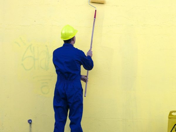 A man paints over graffiti on a yellow wall outdoors.